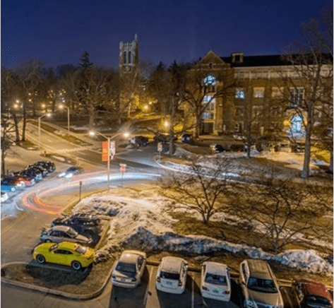 University campus at night with snow-covered grounds and parked cars with light trails visible.