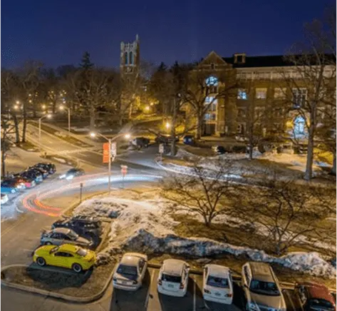 University campus at night with snow-covered grounds and parked cars with light trails visible.