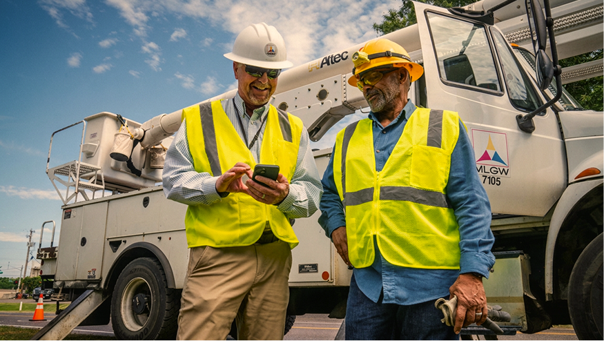 Two MLGW workers in yellow safety vests and hard hats review a smartphone near a utility truck outdoors.