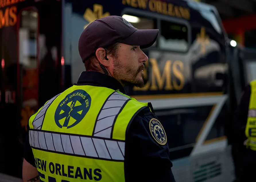 Emergency medical technician in high-visibility vest standing beside New Orleans EMS ambulance at night.