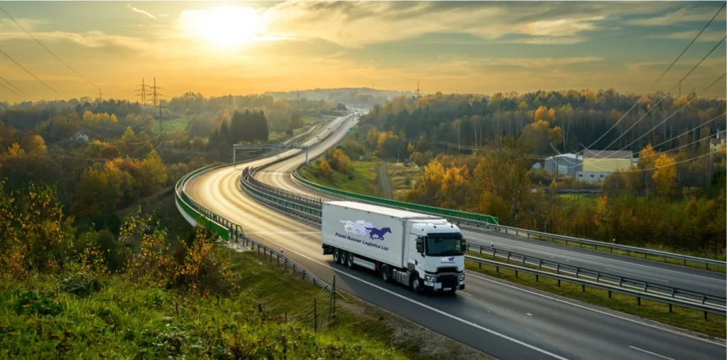 A white Front Runner Logistics Ltd semi-truck drives on a curving highway through autumn foliage under a golden sunset sky.