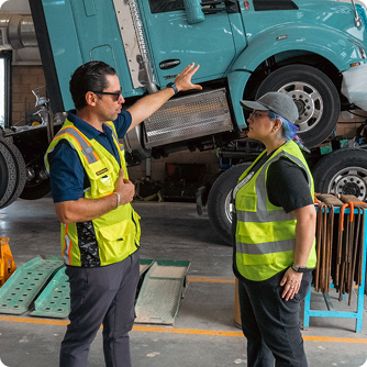 Two workers in safety vests discussing repairs in an auto shop with a turquoise truck on a lift in the background.