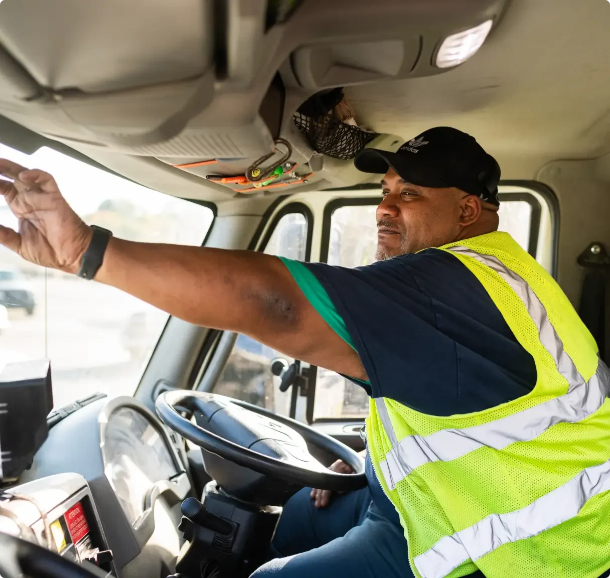 Truck driver in high-visibility safety vest and black cap adjusting side mirror from driver's seat of commercial vehicle.