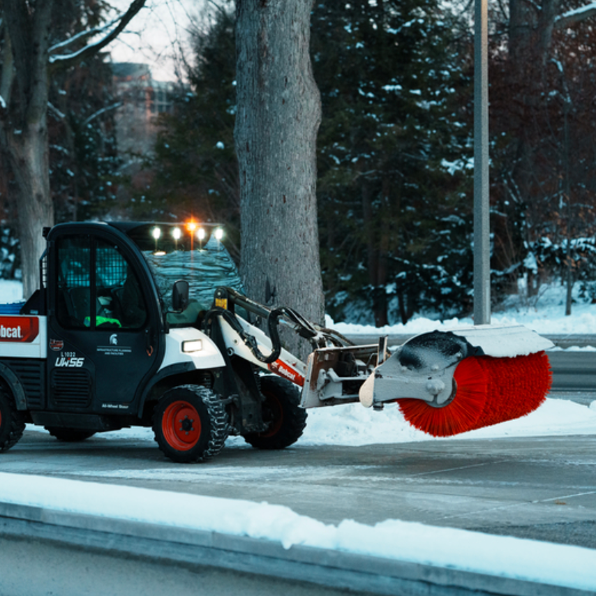 A Bobcat UW56 utility vehicle with a red rotary brush attachment clearing snow from a road in winter.