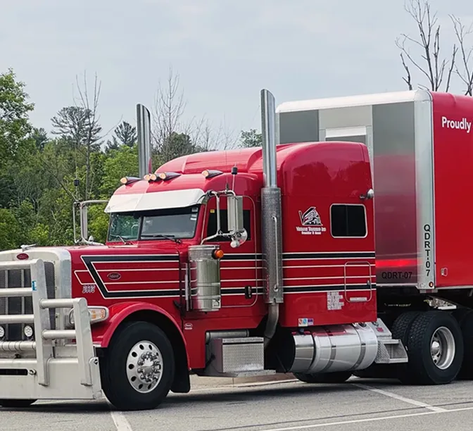 Bright red semi truck with chrome exhaust stacks parked in a lot with trees in the background.