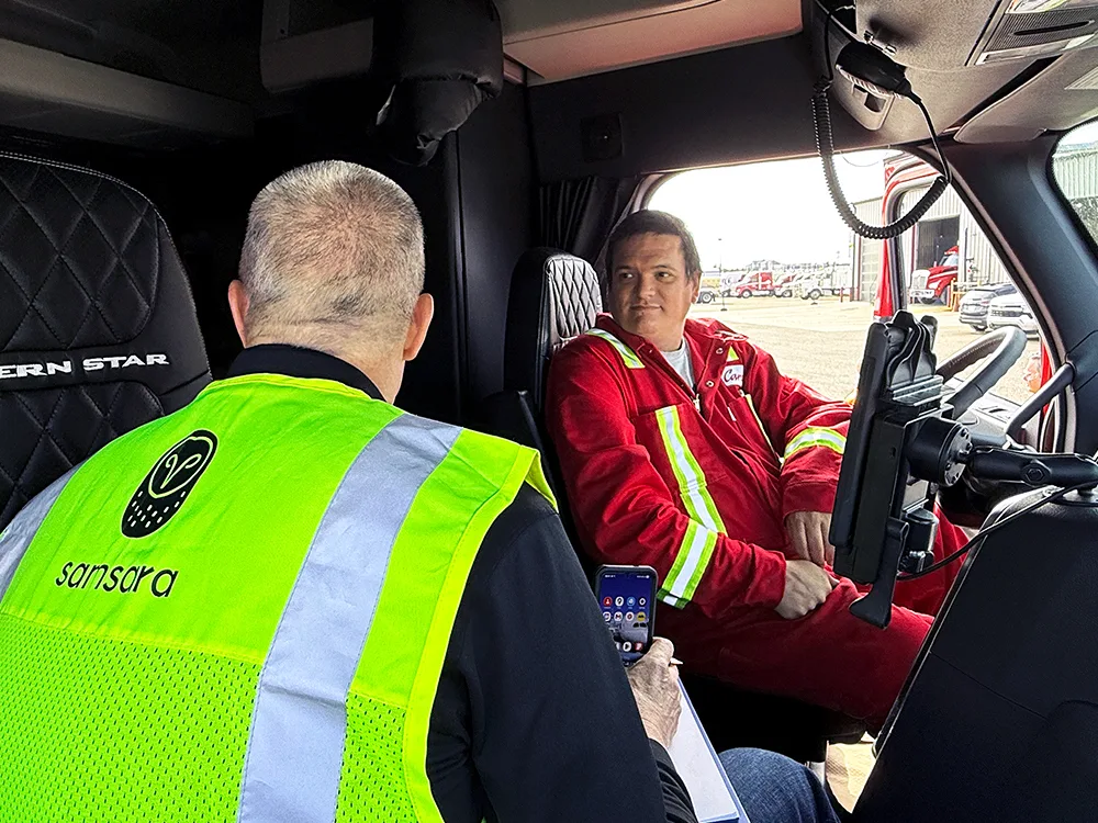 Two workers inside a truck cab - one wearing a neon safety vest, the other in a red uniform with reflective strips.