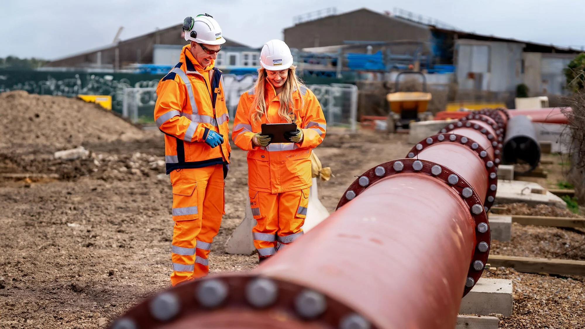 Two construction workers in orange safety gear and white helmets examining a tablet beside a large pipeline at a worksite.