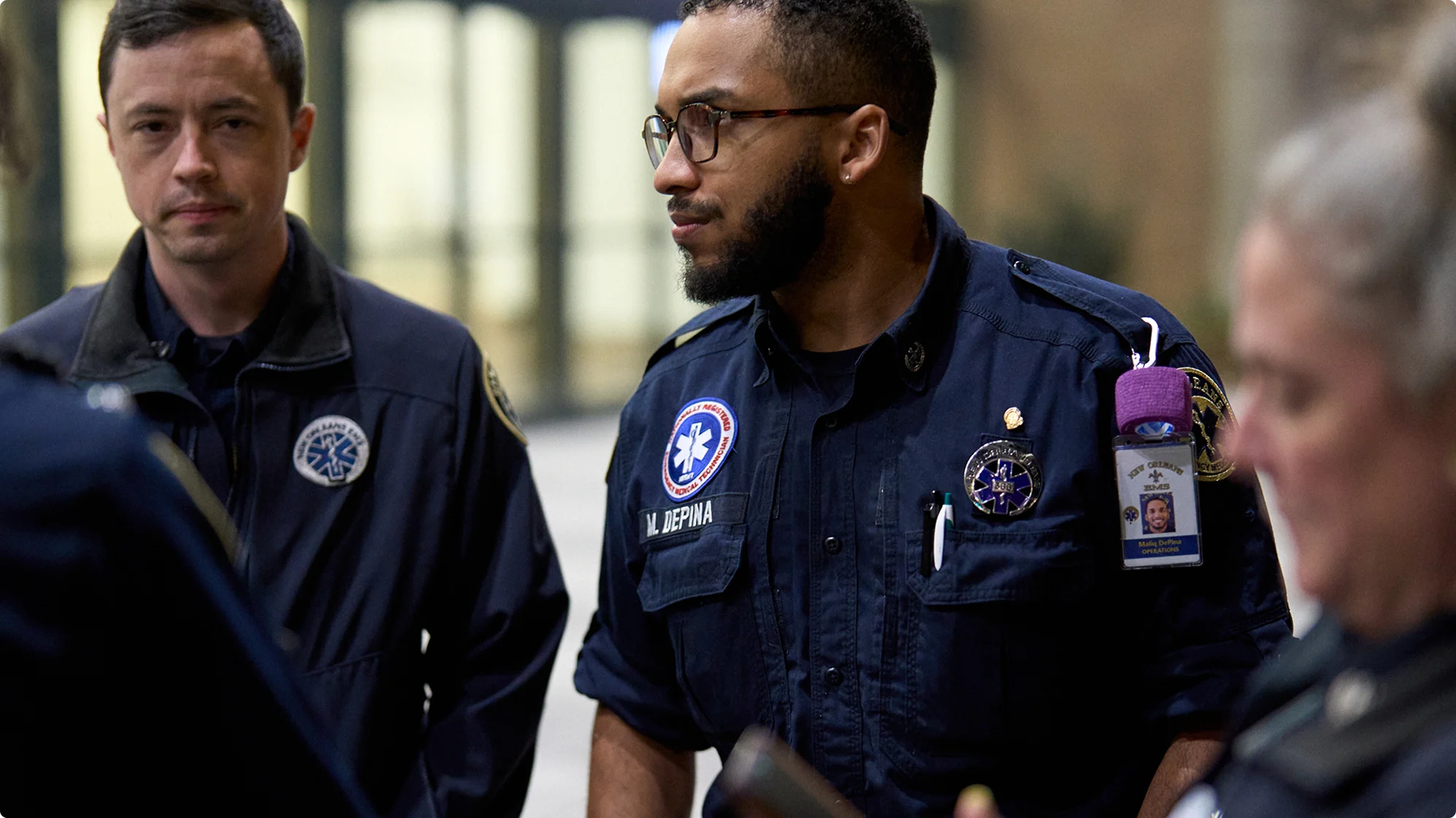Two emergency medical technicians in navy blue uniforms with patches and badges standing together at a scene.