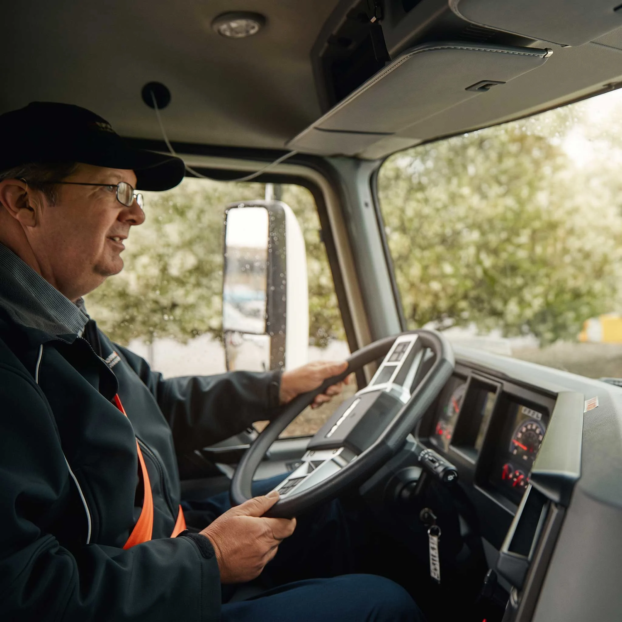 A male truck driver wearing a black cap and glasses sits in a truck cab, holding the steering wheel and looking ahead.