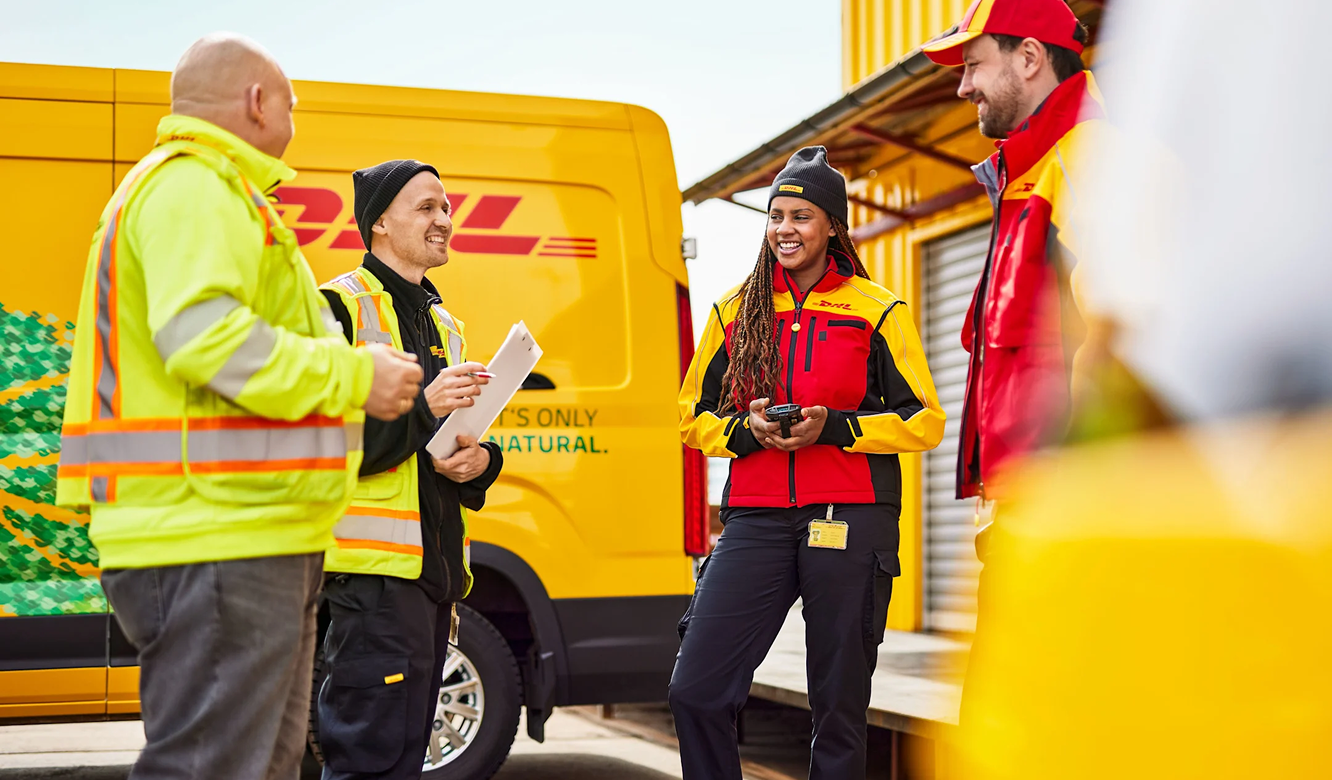Four DHL delivery workers in yellow and red uniforms conversing beside a yellow DHL van at a loading dock.