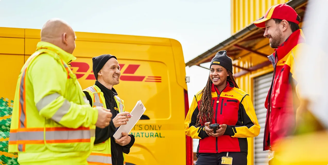 Four DHL delivery workers in yellow and red uniforms conversing beside a yellow DHL van at a loading dock.