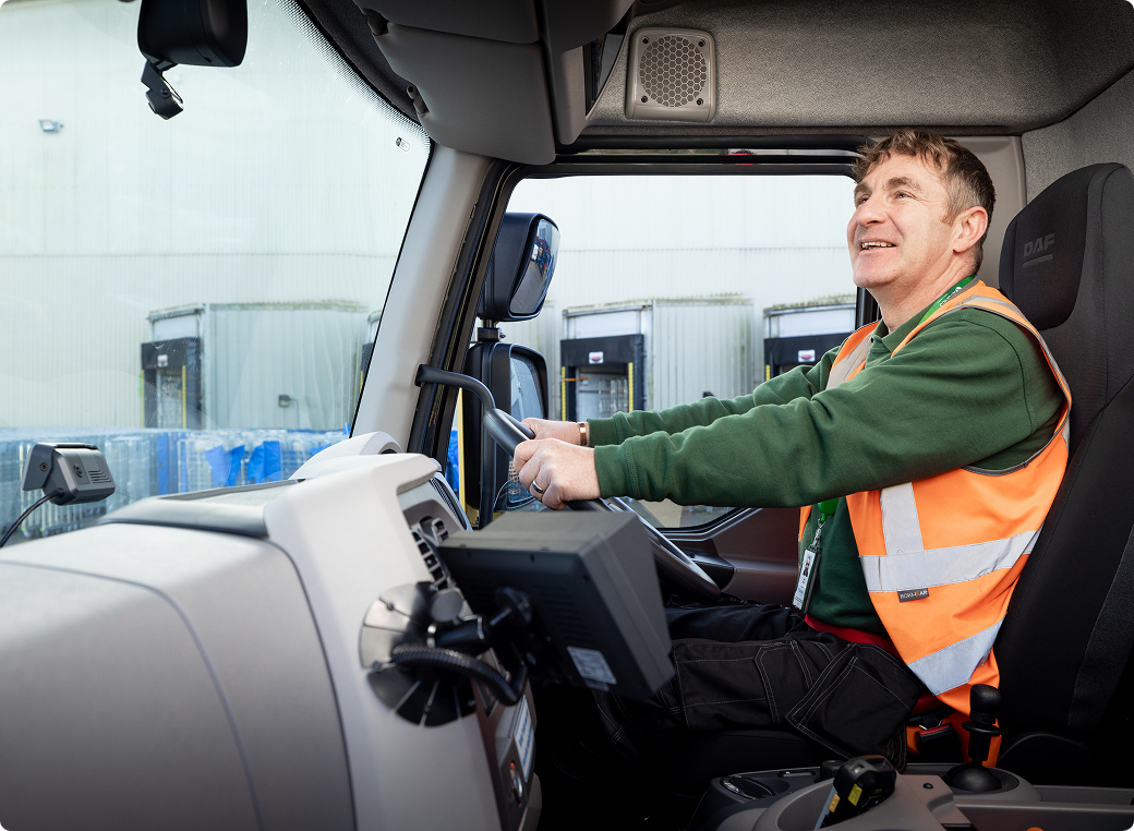 Truck driver in green shirt and orange safety vest smiling while operating a DAF truck at a loading facility.