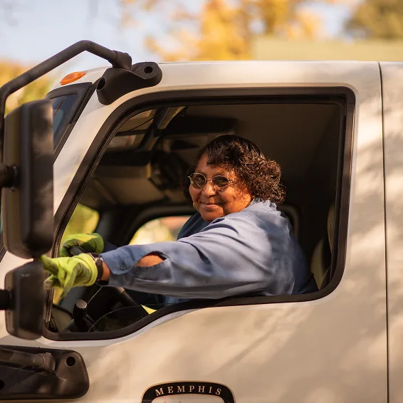 Person in blue uniform driving a white Memphis truck, smiling through the window against autumn background.