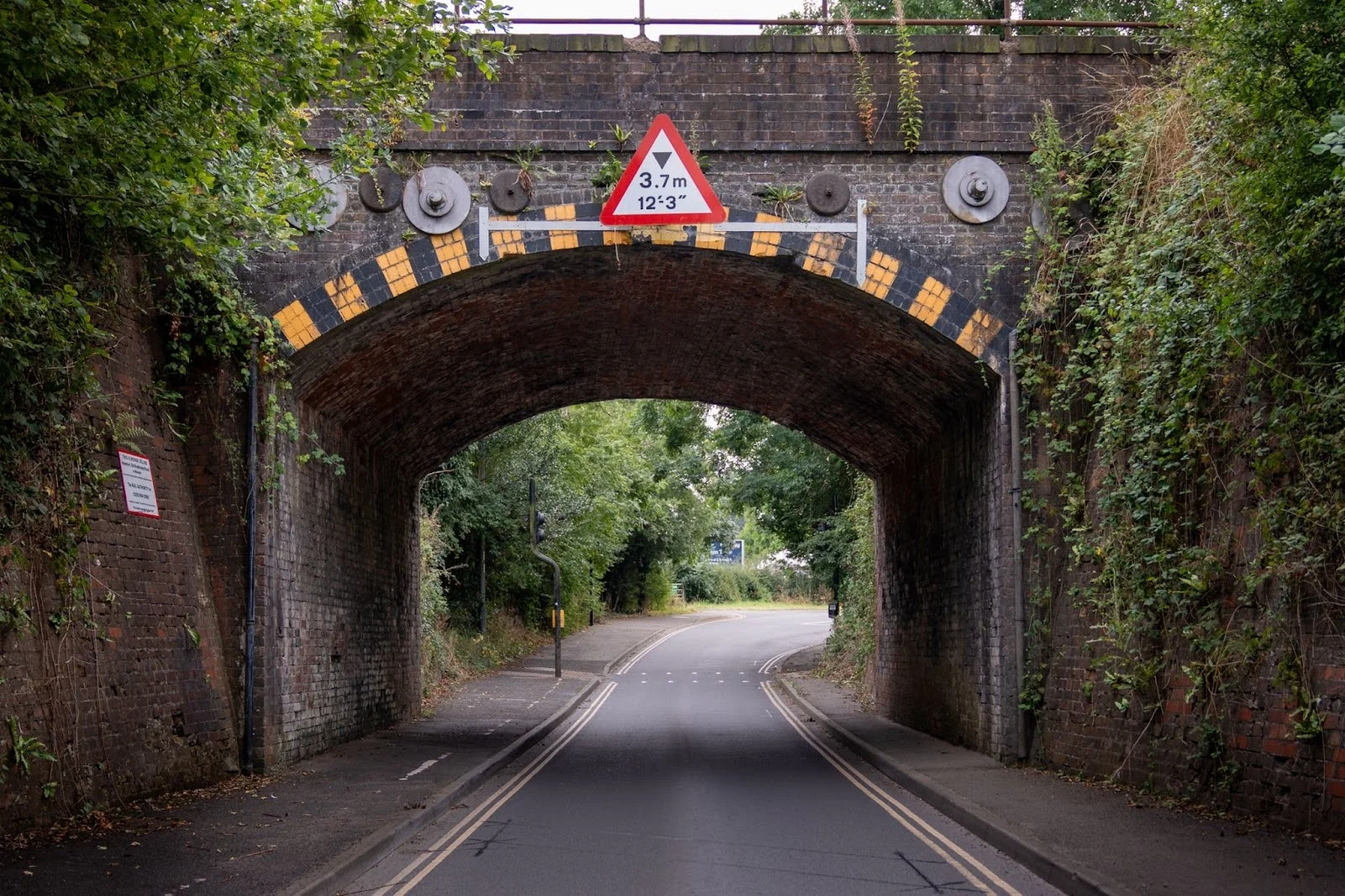 Brick railway bridge over a road with height restriction warning sign of 3.7m, featuring yellow and black markings on the arch.