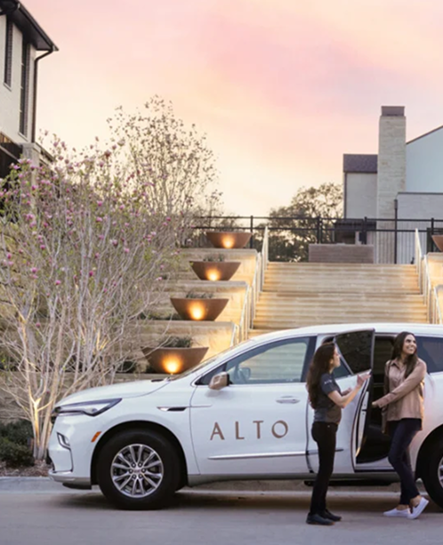 White SUV with "ALTO" logo parked at modern residence with illuminated staircase at sunset