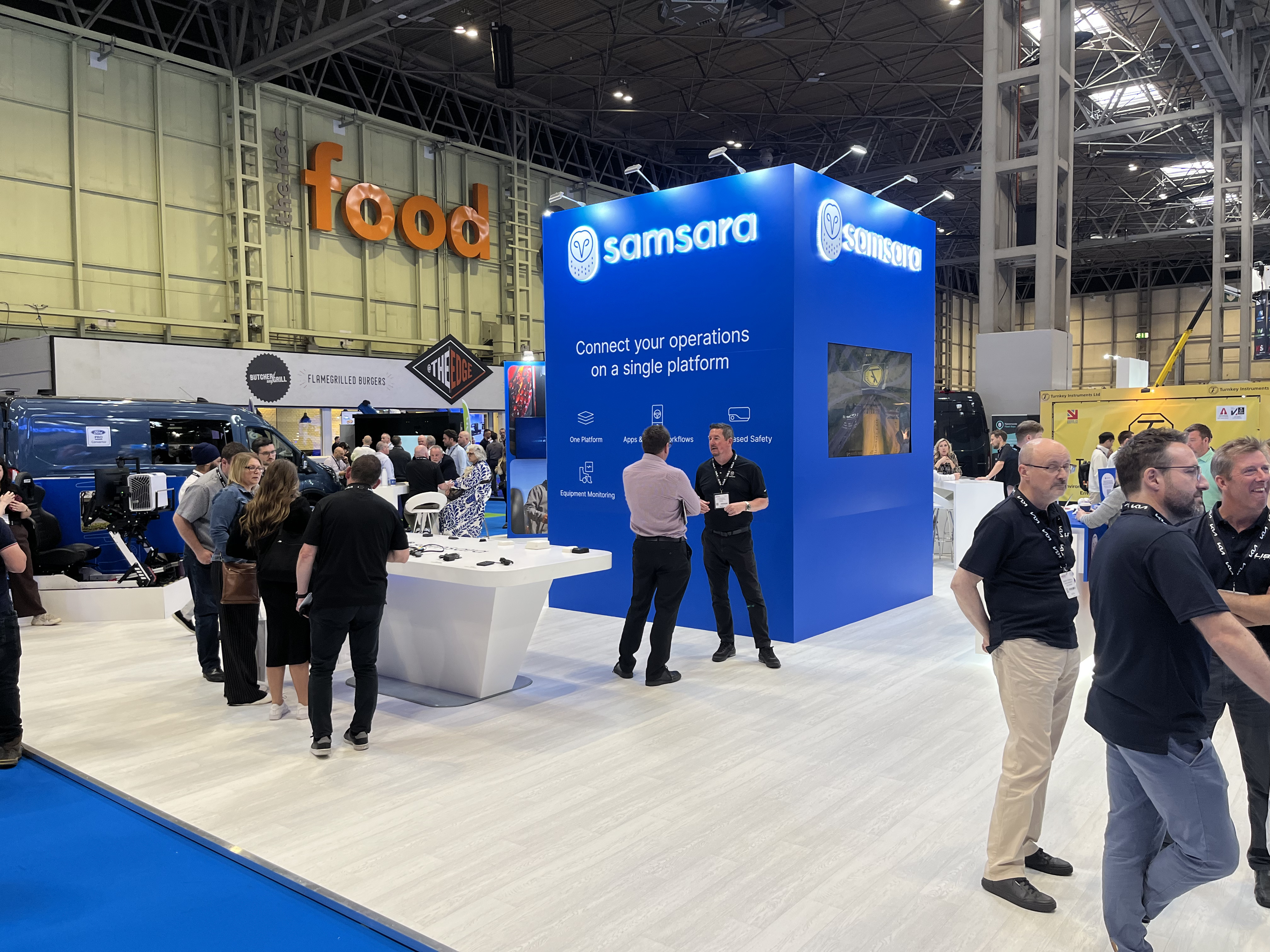 Samsara exhibition booth with blue signage at industry trade show, with attendees networking around white display counters.