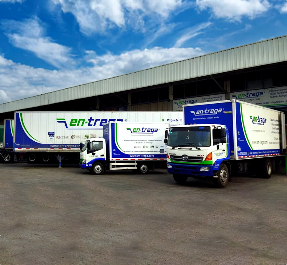 Camiones de reparto de la empresa "en-trega" estacionados frente a un almacén bajo un cielo azul con nubes blancas.