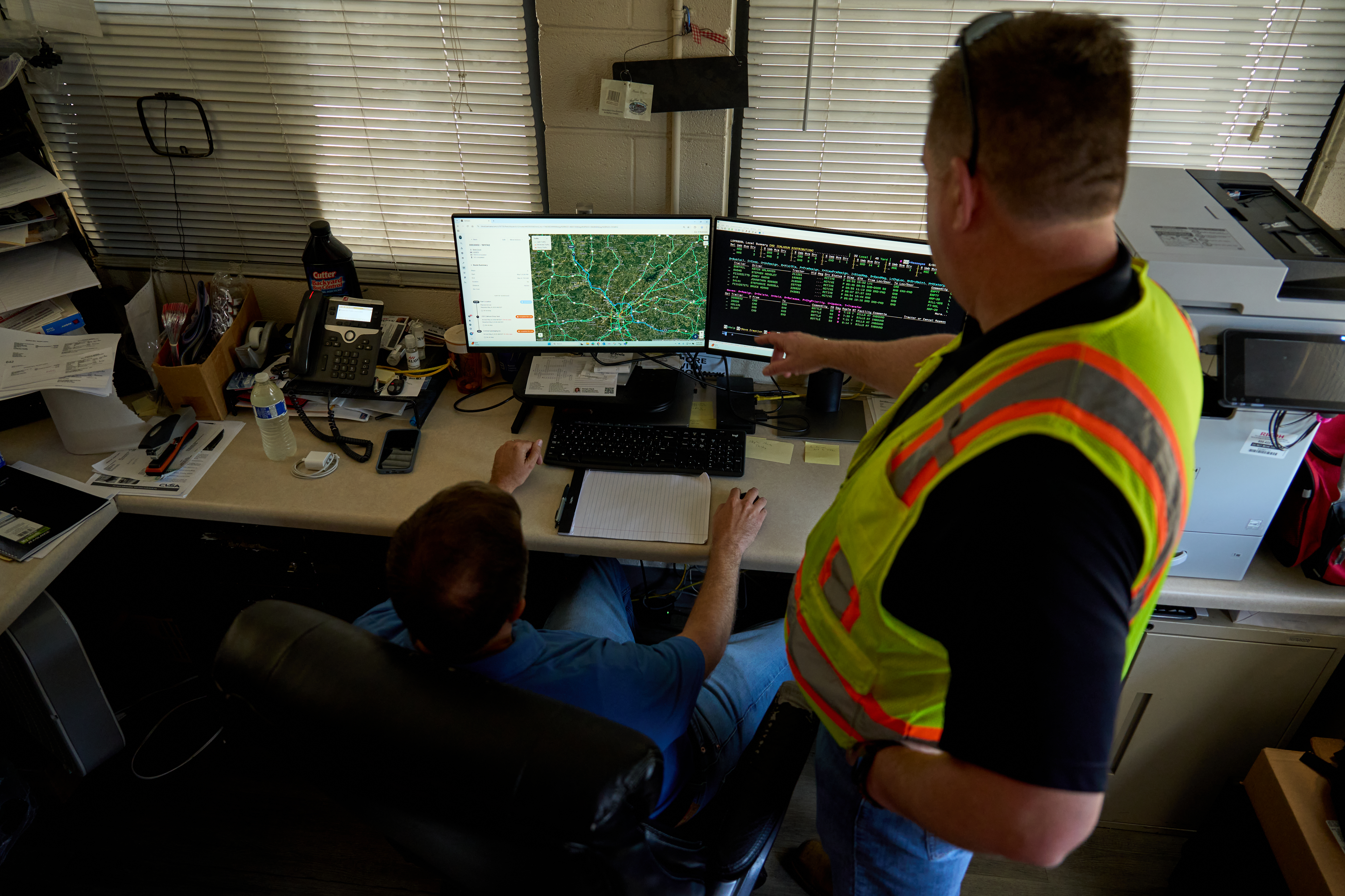 Worker in safety vest pointing at dual monitors showing map and data in an office with window blinds.