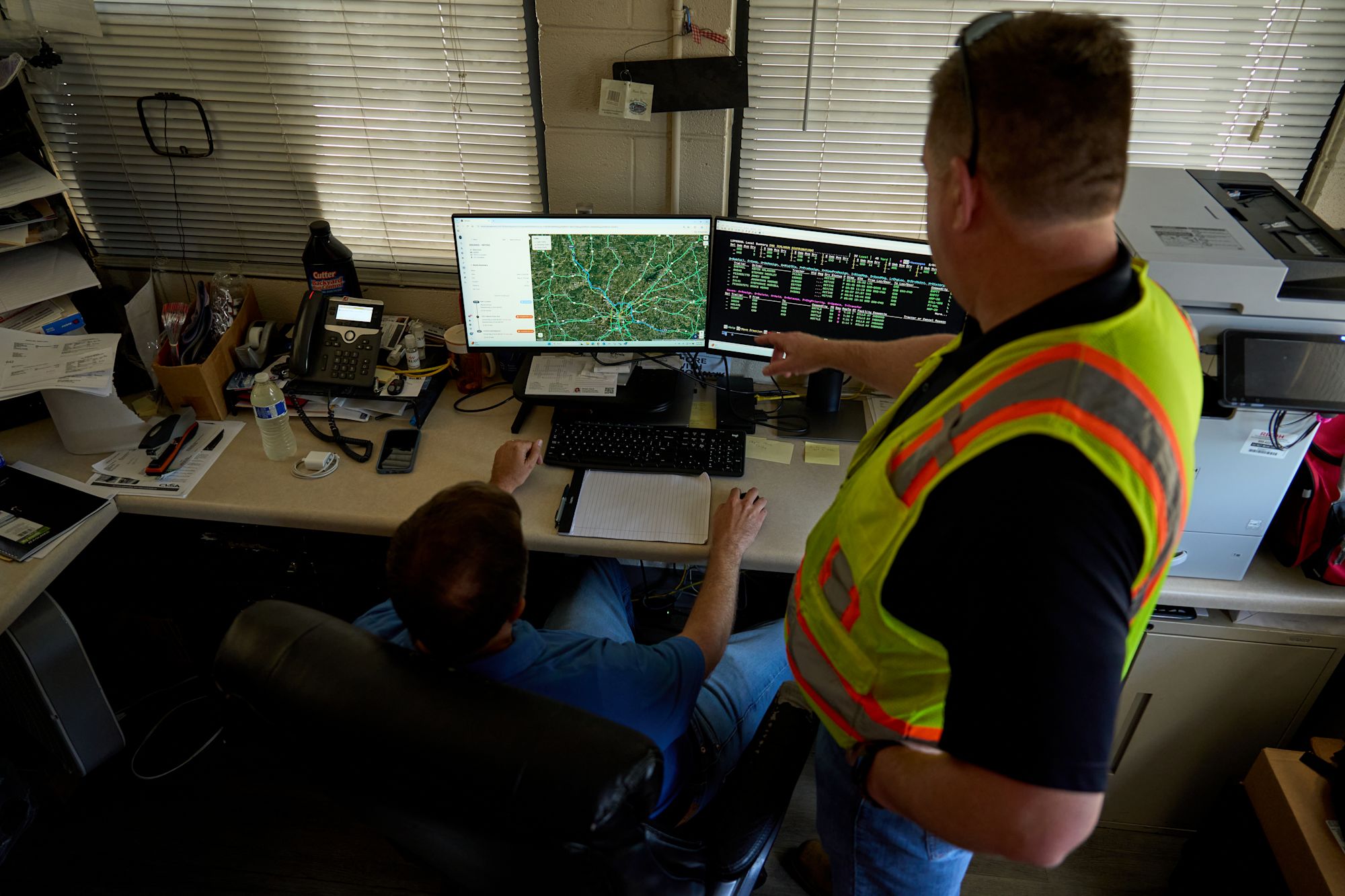 Worker in safety vest pointing at dual monitors showing map and data in an office with window blinds.