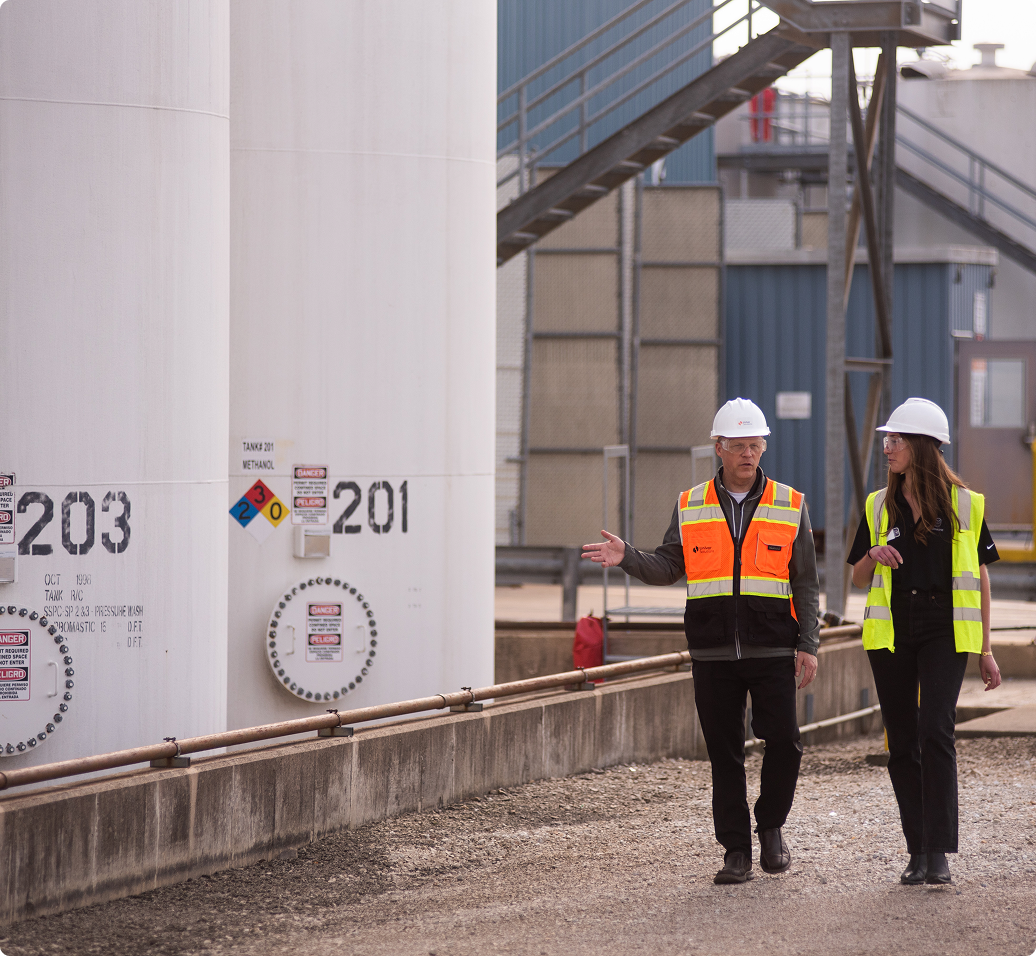 Two workers in safety vests and hard hats walking beside large industrial storage tanks marked with numbers 203 and 201.