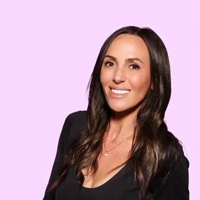 Woman with long dark hair smiling against a light pink background, wearing a black top and delicate necklace.