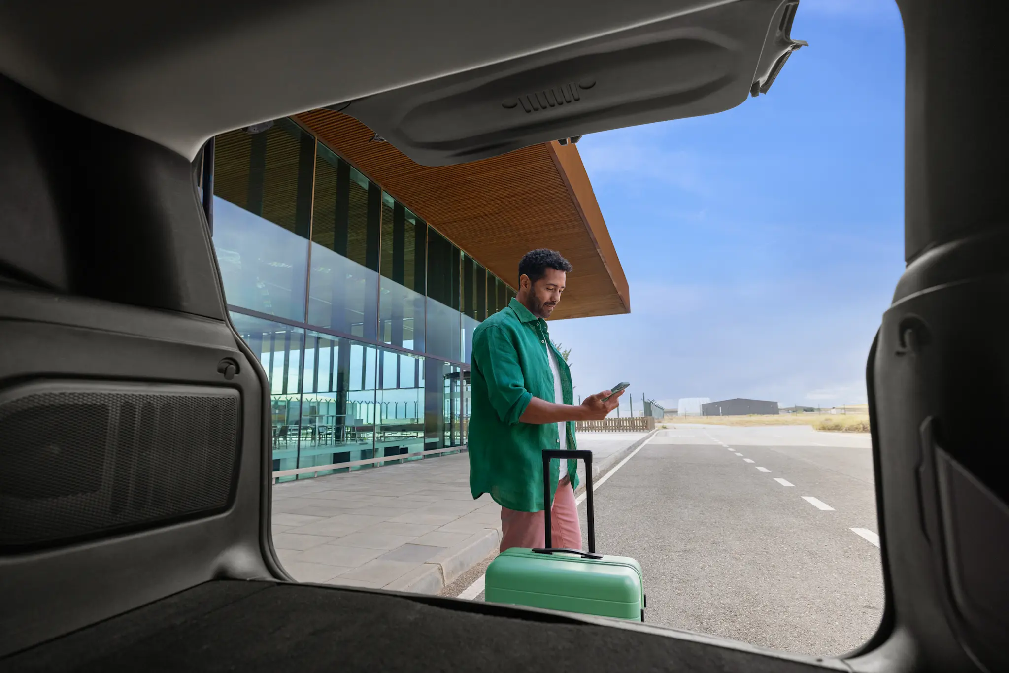Free2move carsharing customer with a green suitcase standing outside an airport, checking their phone, viewed from the open trunk of a car.
