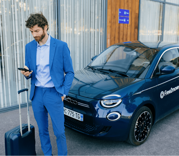 Client de l'autopartage Free2move avec une valise verte debout devant un aéroport, regardant son téléphone, vu depuis le coffre ouvert d'une voiture.