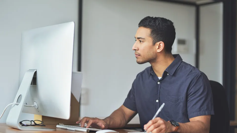 A professional typing at a desk, taking notes and looking at his monitor.