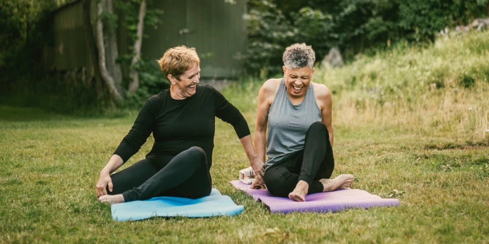Deux femmes sur des tapis de yoga à l’extérieur en riant et en faisant de l’exercice