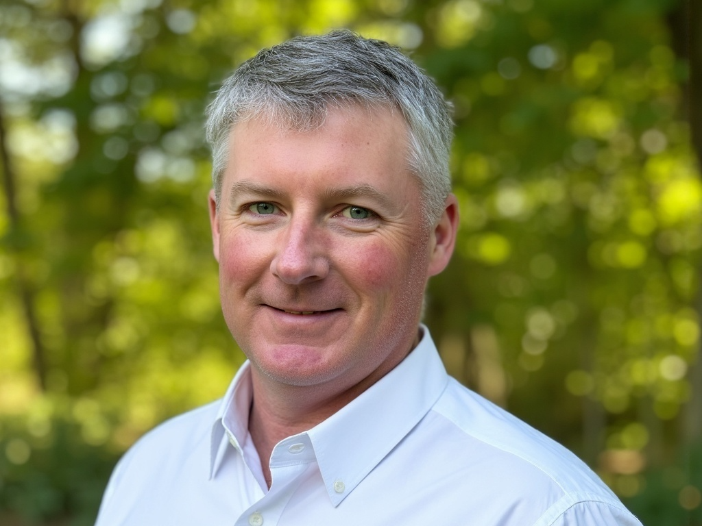 Josh Stewart, a white male with grey and white hair dressed in a white button up with dappled sunlit trees in the background
