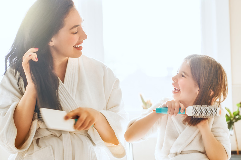 A woman and her little daughter are in white bathrobes, brushing their hair after a shower.