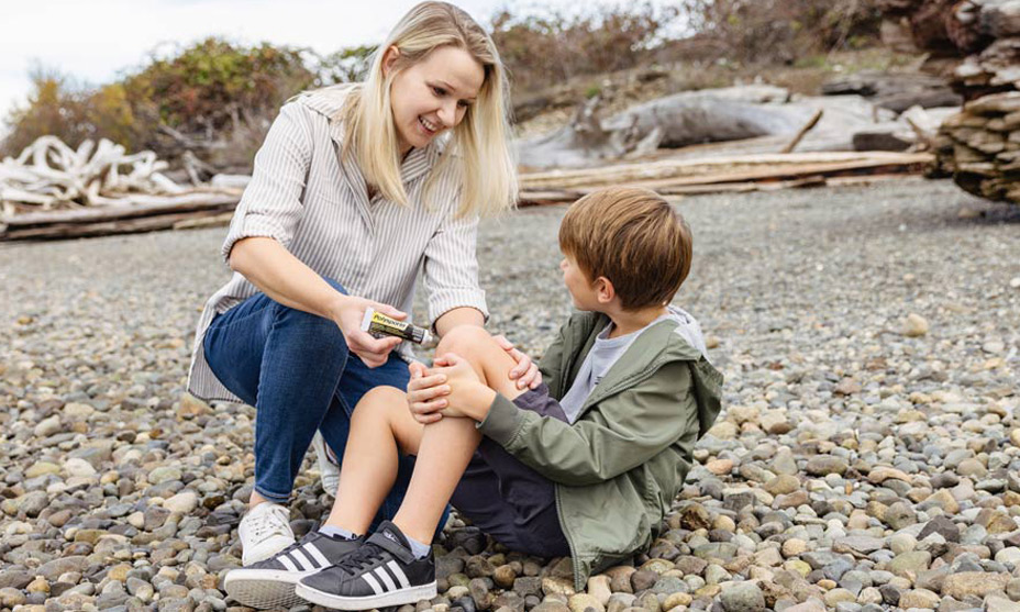 Young boy sitting on a pebble beach with his jacket on and his mother applying ointment on his knee while smiling.