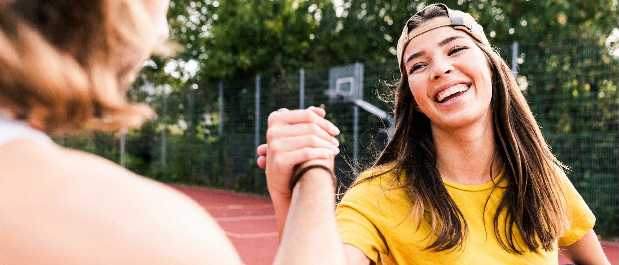 Jeune femme souriante portant un chapeau et tenant un ballon de basketball qui serre la main d’une amie sur le terrain de basketball extérieur | LISTERINE