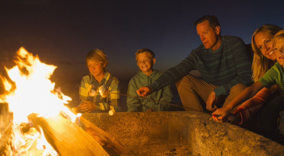 Jeunes parents et leurs trois petits garçons préparant des choco-guimauves autour d’un feu de camp.