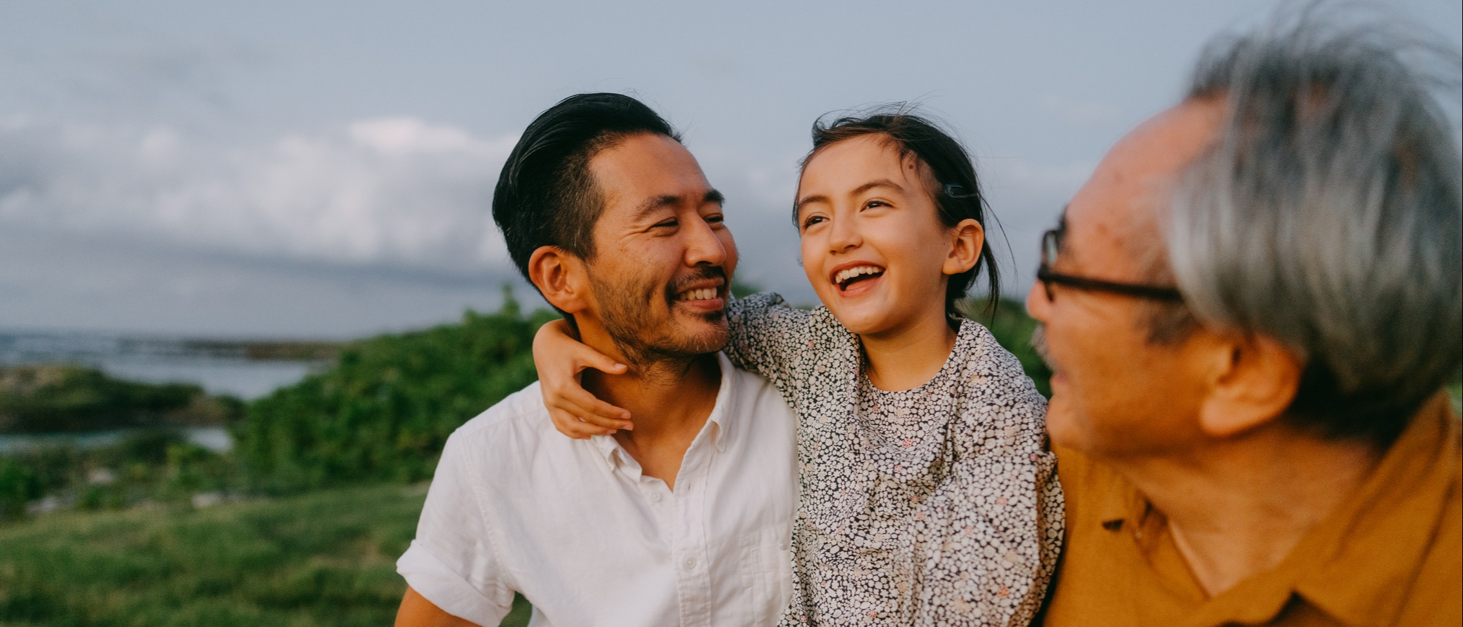 Young girl sitting on her father's lap next to her grandfather while smiling outdoors