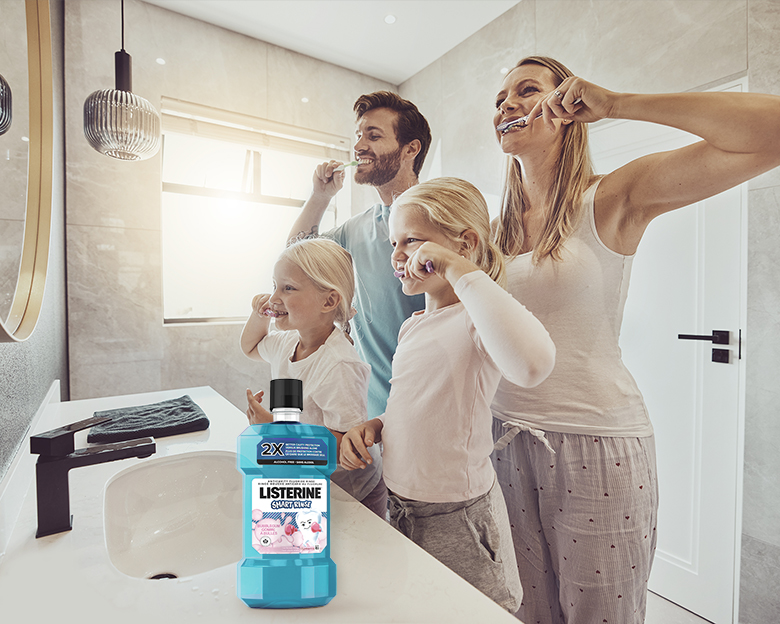 Young couple with their two little girls looking at the bathroom mirror and brushing their teeth while a Listerine Mouthwash bottle is on the counter