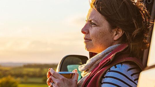 Woman with a scarf around her neck leaning on her car and holding a cup of hot beverage on the side of a country road.