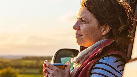 Woman with a scarf around her neck leaning on her car and holding a cup of hot beverage on the side of a country road.