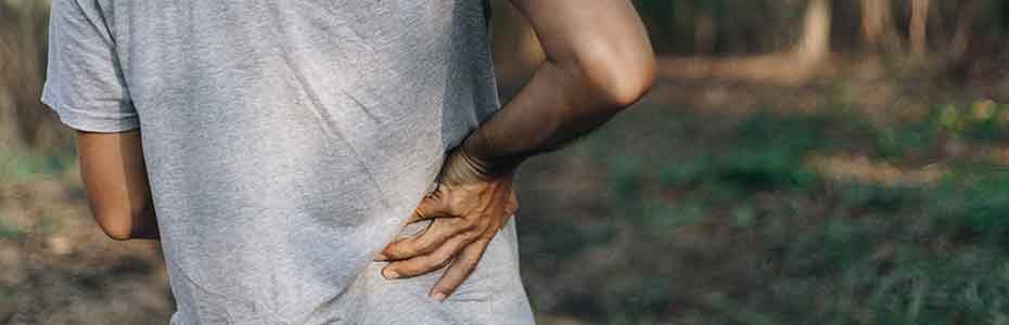 Man experiencing back pain while walking outdoors and holding his lower back with his right hand<br />
