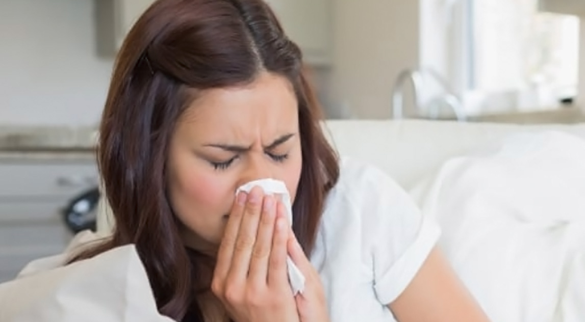 Sick woman lying on a couch under a comforter and blowing her nose into a paper tissue