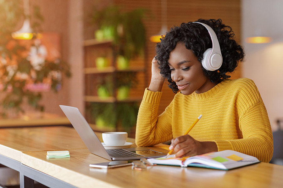 A young woman is sitting in front of her laptop, taking notes, and wearing a headset.