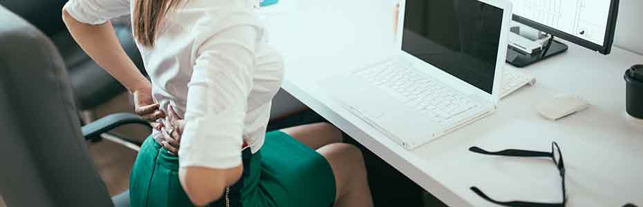 Woman who is sitting on a desk working on her computer experiencing pain and holding her lower back with her hands<br />
