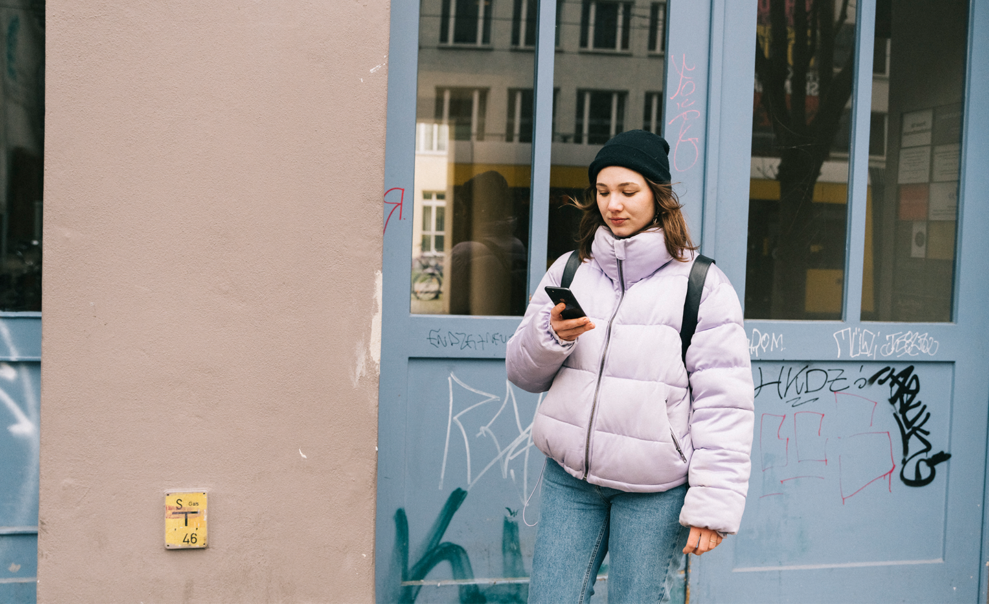 This image depicts a medium closeup of a woman standing with her back to a building, reading something on her mobile phone.
