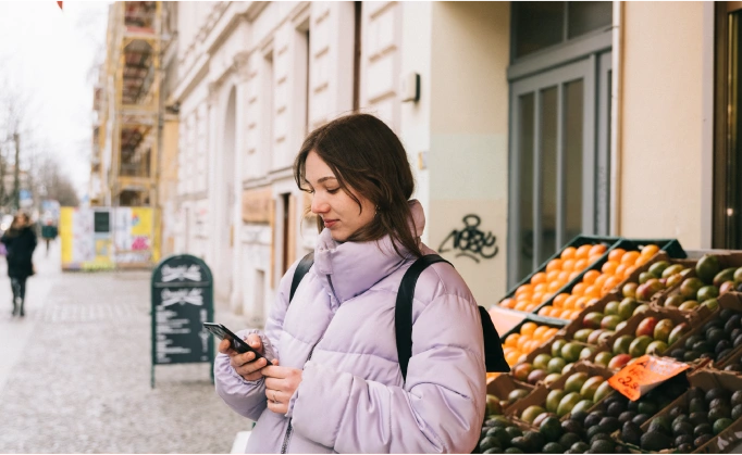This image depicts a woman standing in a streetscape, looking at her mobile phone.