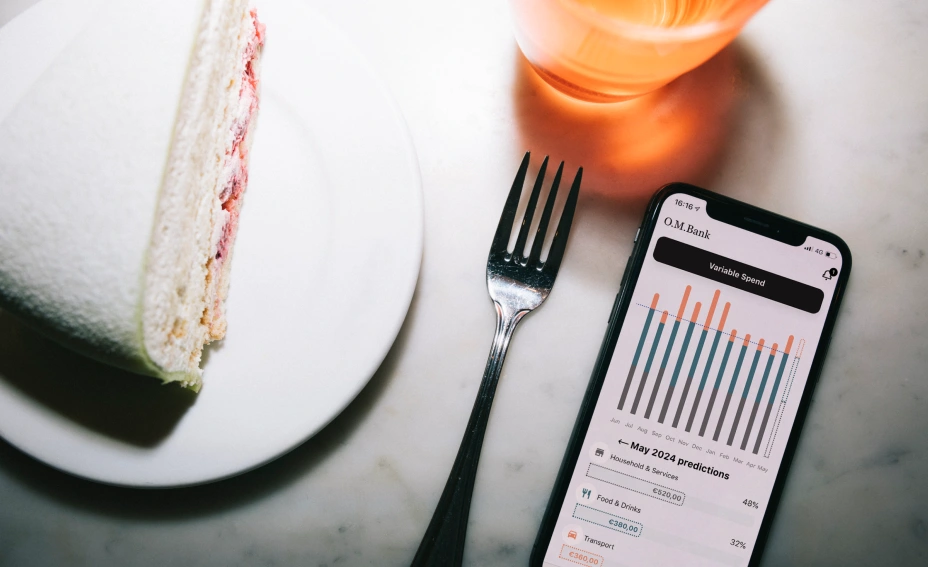 This image depicts a tablescape from above with a slice of cake, fork, drinking glass and mobile phone, with the phone screen displaying Tink's Variable Spend feature in action.