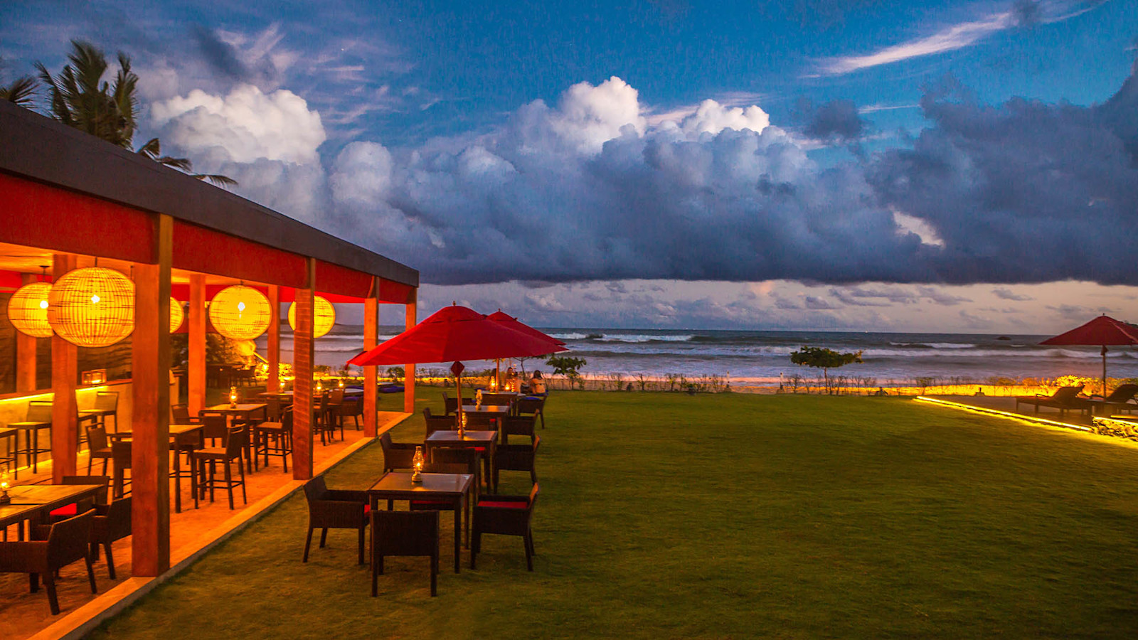 The beach restaurant at dusk: lit tables under round paper lanterns in an open pavilion, more tables on the lawn under red umbrellas, and the sea beyond