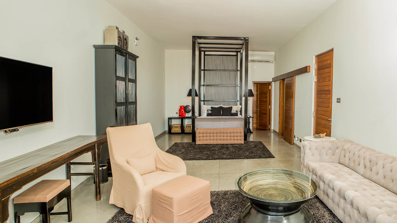 The Executive Suite: a long room with a four-poster bed at the far end, a tufted white sofa, an armchair with a footstool, a writing desk and a wall-mounted TV
