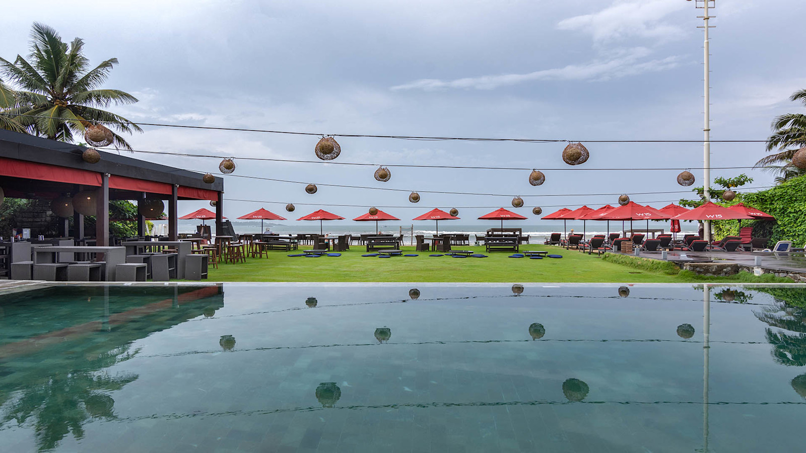 The pool in front of the lawn under a grey sky, with woven lanterns strung overhead, red umbrellas and an open-sided restaurant along the beach