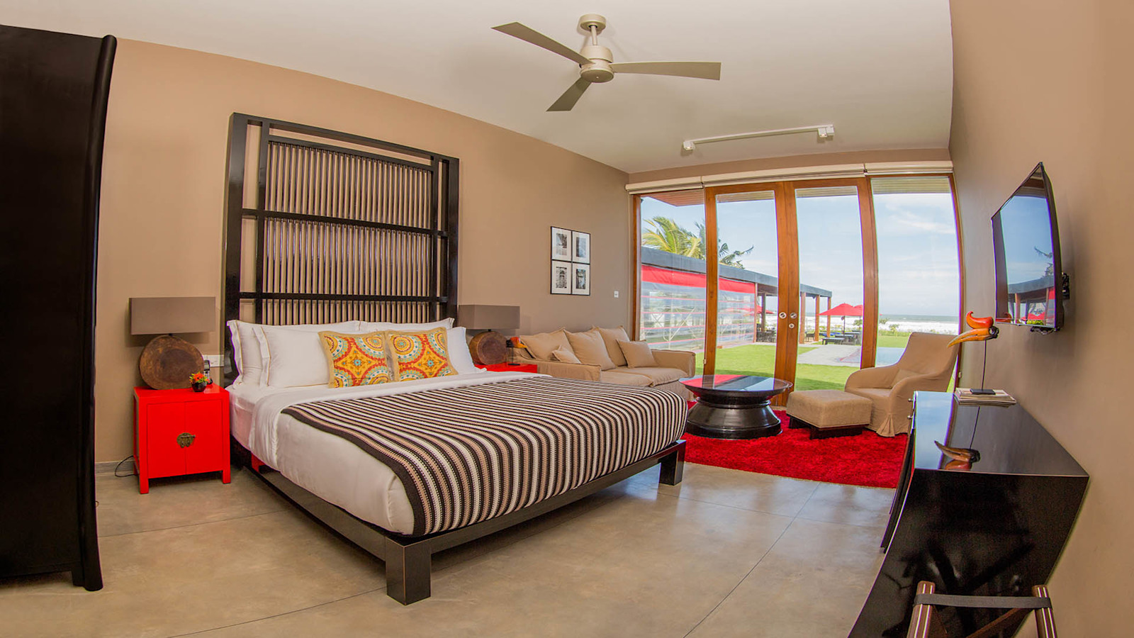 An Ocean View Deluxe room: a low double bed with a striped throw, a red bedside cabinet, a sofa and armchair, and full-height glass doors looking over the lawn to the sea