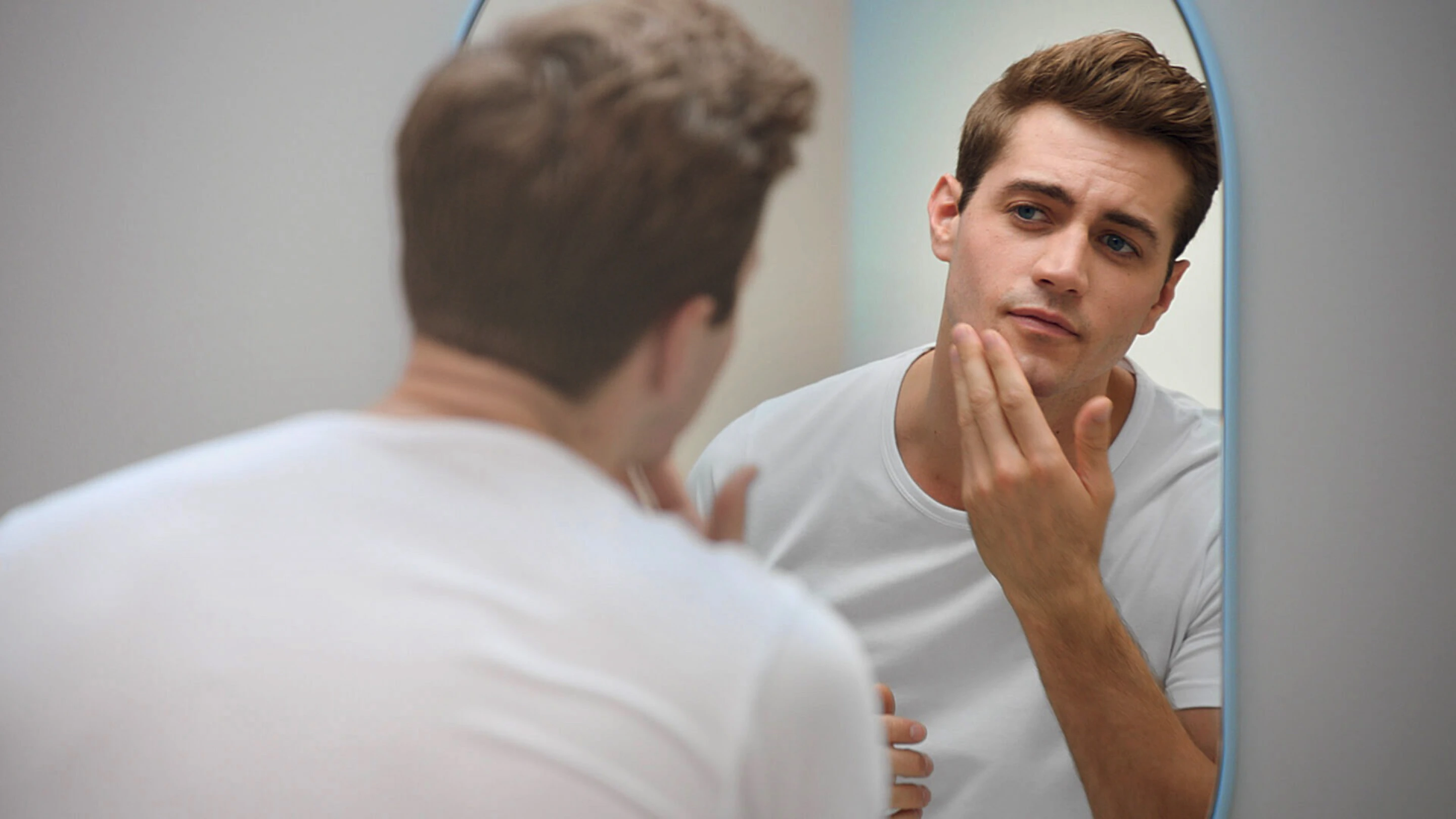 Man is looking at himself in the mirror, his silhouette is blurred while the mirror image is clear. He seems content and is about to touch his face.